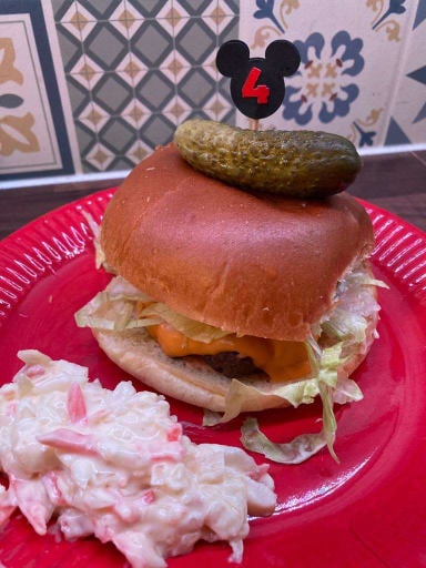 One of the gourmet burgers made by a LDSA member. The burger is placed on a red plate alongside some homemade coleslaw and features a gherkin held in place by a skewer. 