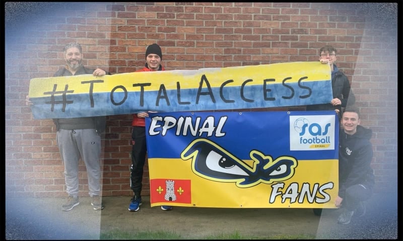 four fans holding up a couple of blue and yellow banners with #TotalAccess on one and SAS Epinal Fans on the other. They are all looking at the camera and smiling.