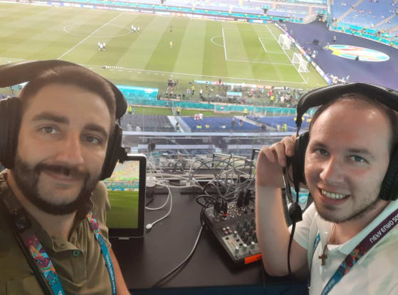 two audio-descriptive commentators sitting in the press box of a stadium at the Euros, both wearing their headsets.